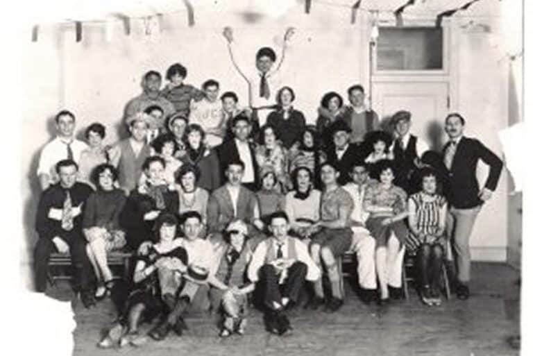 A black and white photo of a large group of people posing together indoors, some sitting and others standing.