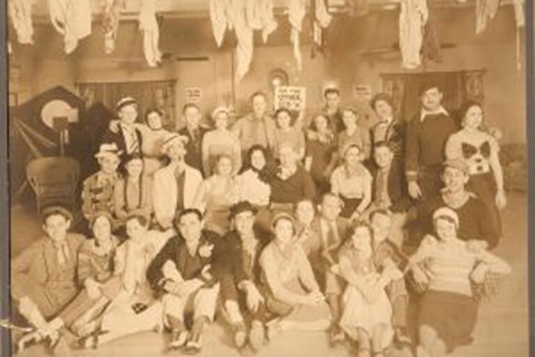 A sepia-toned photo of a large group of people posing indoors, with decorations hanging from the ceiling.