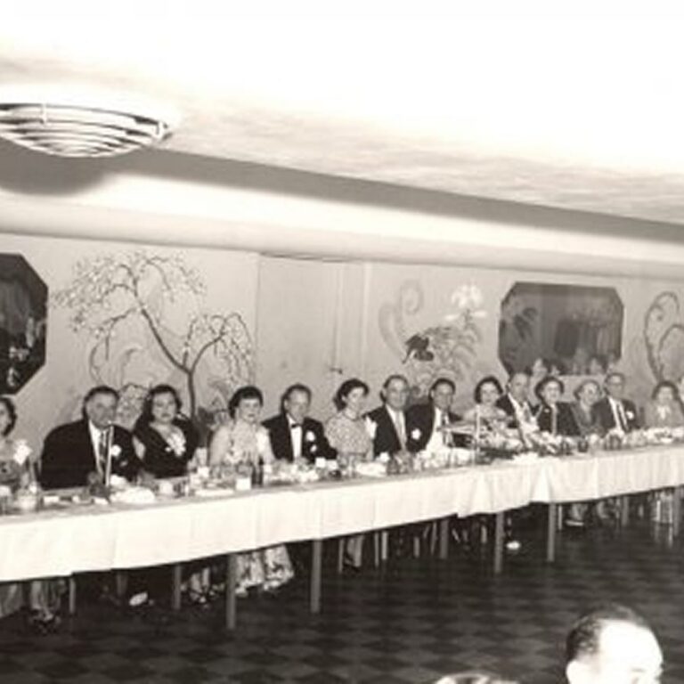 A group of people in formal attire sit at a long banquet table in a decorated room, smiling for a photo.