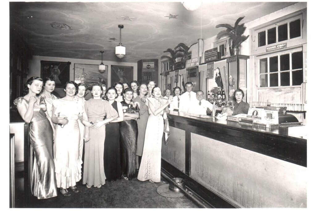 A group of women in formal dresses stand near a bar counter with staff behind it in a vintage setting.