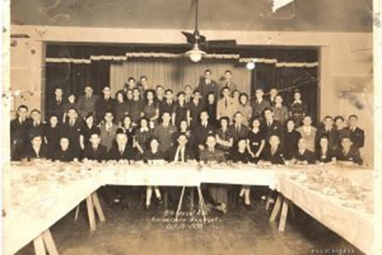 A large group of people pose behind banquet tables in a hall, with a stage and curtains in the background.