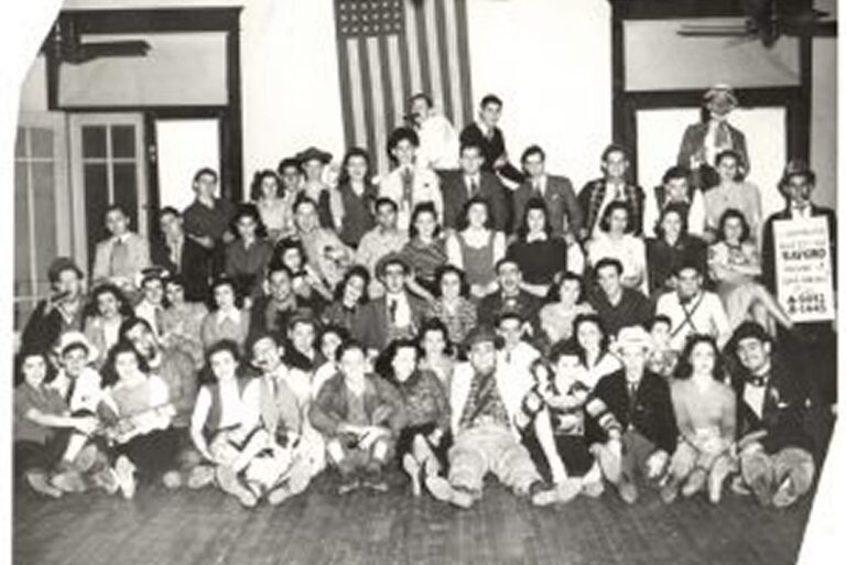 Large group of people in costumes pose indoors in front of a large U.S. flag, smiling at the camera.
