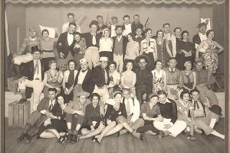 Large group of people in costumes posing on a stage, vintage sepia photo, some seated and some standing, smiling.