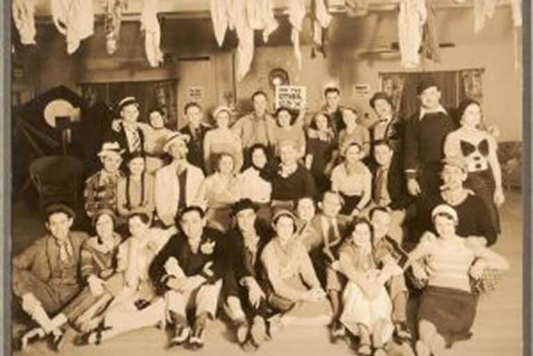 Vintage photo of a large group of people posing indoors with decorations hanging from the ceiling above them.