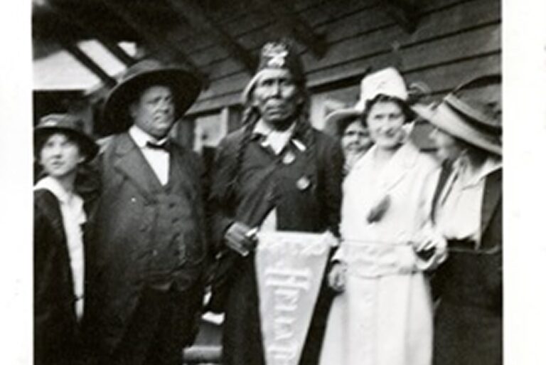 Five people, including a man in native dress holding a pennant, pose together in front of a wooden building.