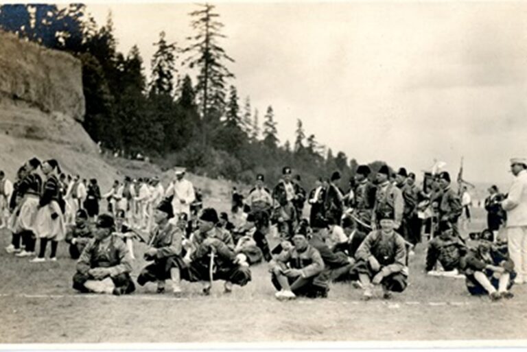 A group of men in traditional clothing gather outdoors on a field, with trees and a cliff in the background.