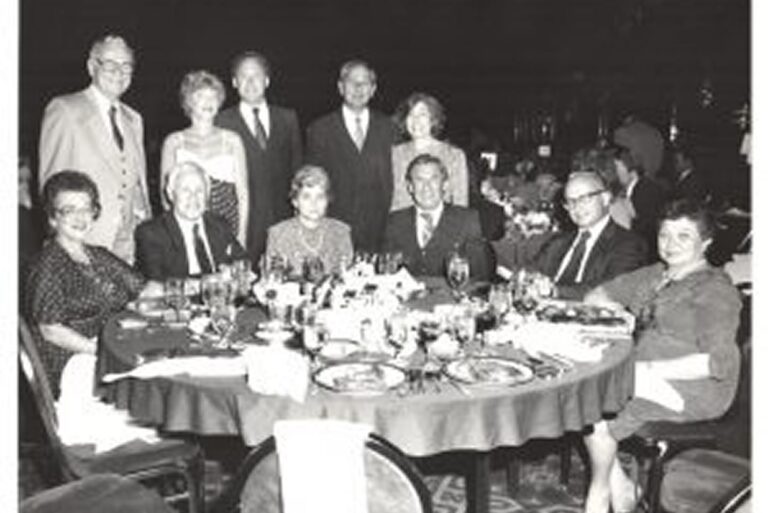 Ten people, six seated and four standing, pose for a group photo around a formal dinner table at an event.