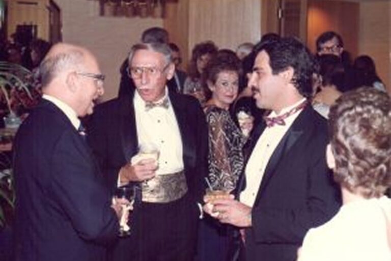 Three men in tuxedos talk and hold drinks at a formal indoor party, with other guests mingling in the background.