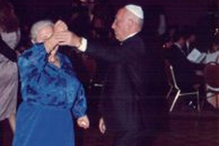 Elderly couple dancing together; man wears a white kippah and suit, woman wears a blue dress.