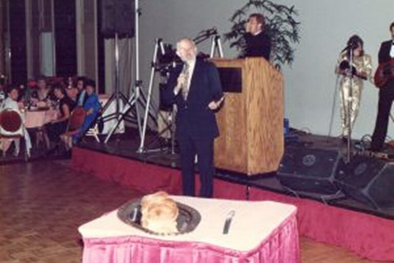 A man speaks on stage as a woman’s head appears on a table with a knife and fork beside it, creating an illusion.