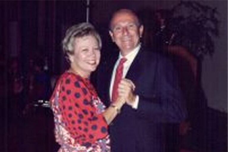 An older couple smiles while dancing together at an indoor event. The woman wears red, the man wears a suit and tie.
