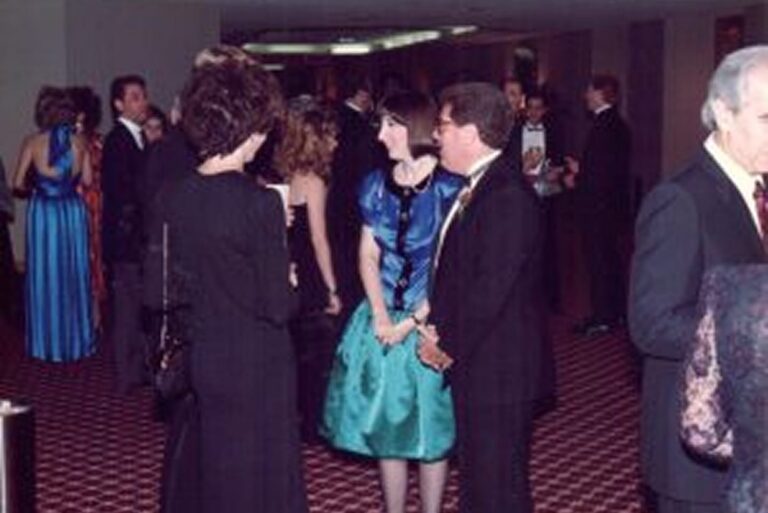 People in formal attire converse in a carpeted hall at an indoor event; some face the camera, others are turned away.