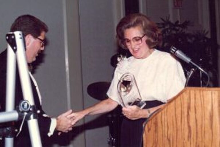 Woman holding an award shakes hands with man at podium, both smiling during a formal event.