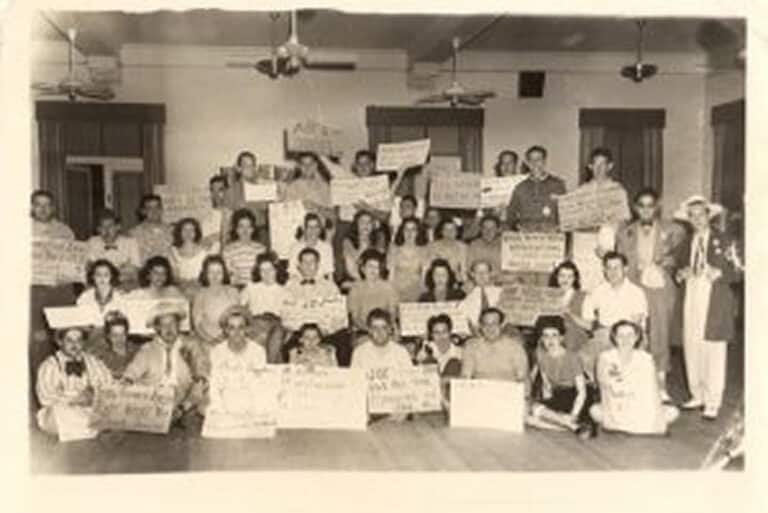 A large group of people pose indoors, holding handwritten signs, in a black and white vintage photograph.