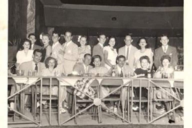 A group of men and women in formal attire sit and stand around a long table in a vintage setting.