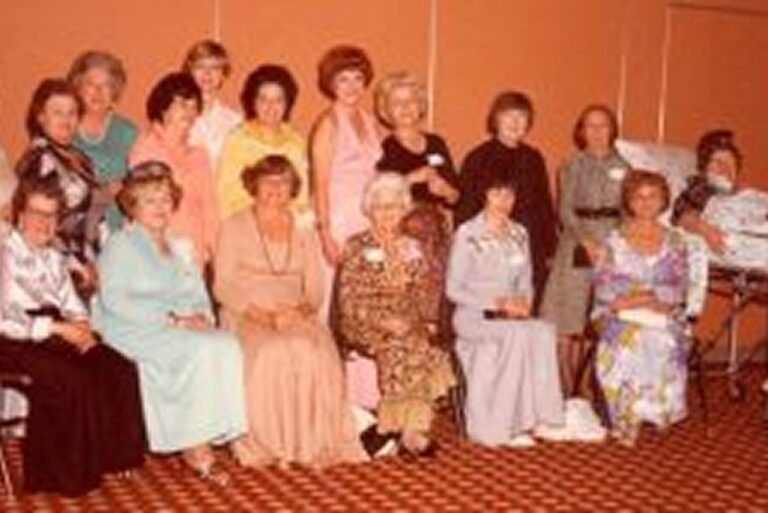 A group of women in formal dresses pose together indoors on patterned carpet in front of orange walls.
