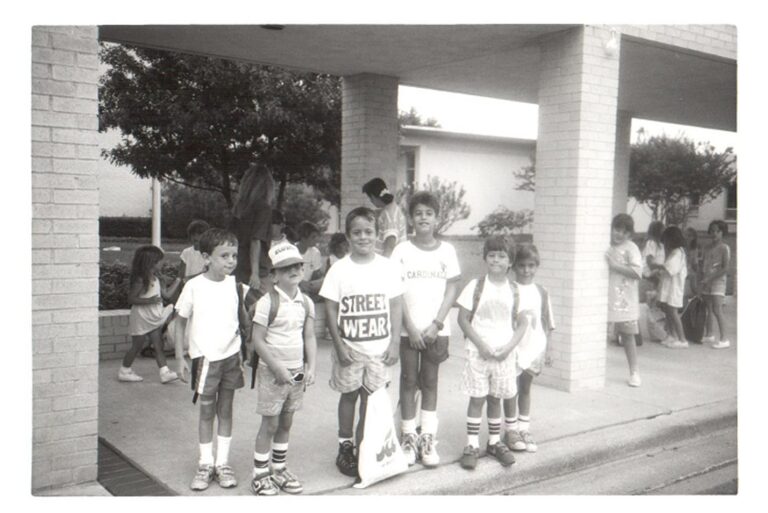 Five boys stand together outside a school building, with more children and adults gathered in the background.