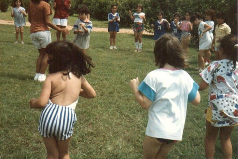 Children stand in a circle on grass, playing a game outdoors on a sunny day.
