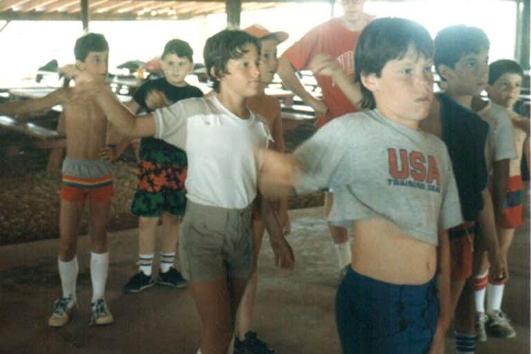 A group of young boys exercise indoors, some with arms raised. One boy’s T-shirt says “USA Training Day.”.