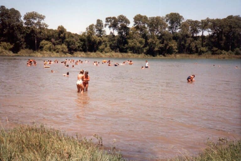 People swimming and wading in a muddy river, with trees lining the opposite bank under a clear sky.