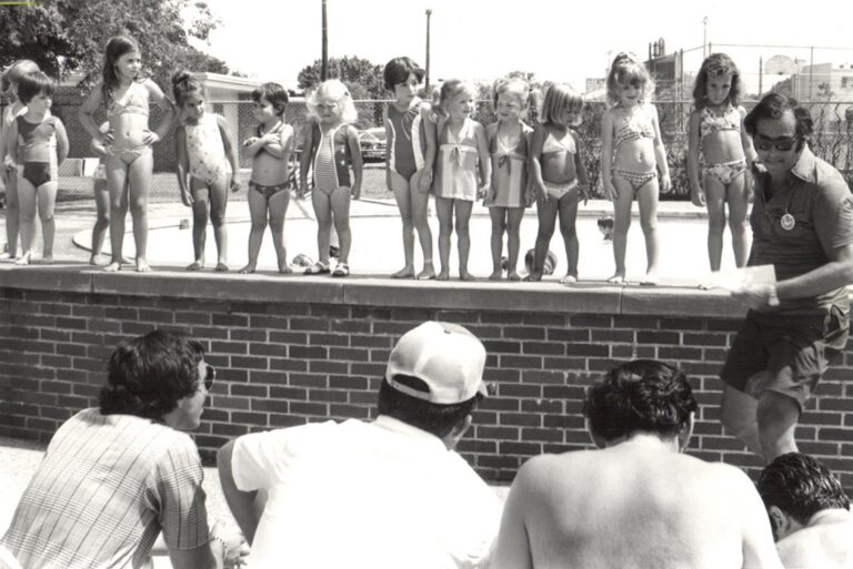 A group of young children in swimsuits stand in a line by a pool as adults watch and a man kneels in front of them.