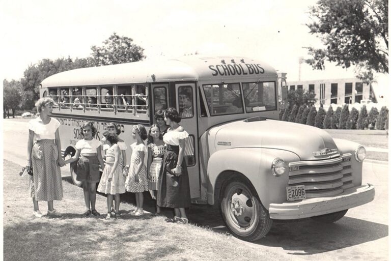 1950s school bus parked with a woman and six girls standing beside it, trees and a building in the background.