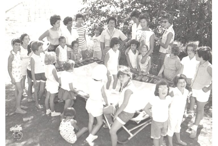 A group of children and adults gather outdoors around a table, smiling and talking on a sunny day.