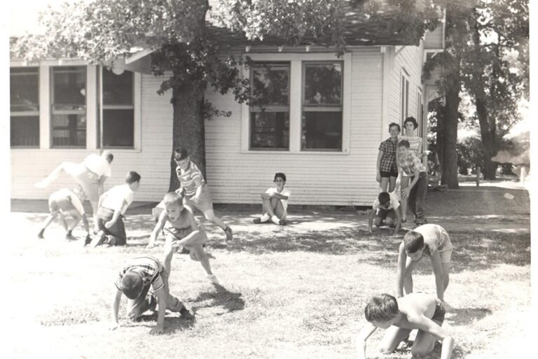 Children play leapfrog on grass near a house, while others watch from the shade and sit by the wall.