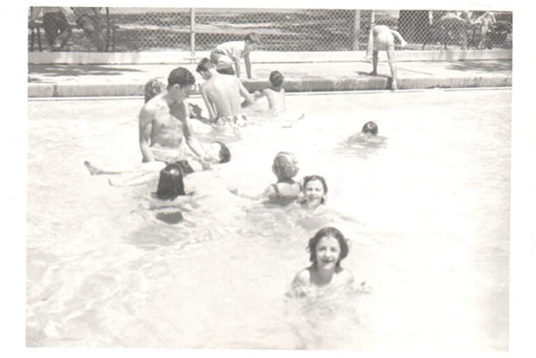 Children swim and play in an outdoor pool on a sunny day, with some people sitting and standing by the poolside.