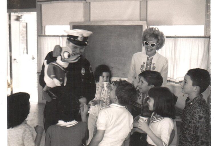 A police officer with a dog puppet entertaining smiling children and a woman in sunglasses at a classroom gathering.