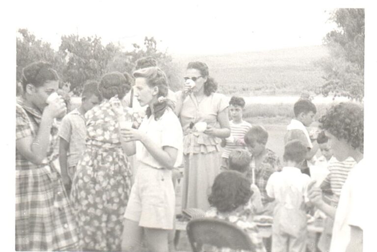 Vintage photo of kids and adults outdoors, eating and talking near a table on a sunny day.
