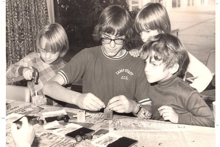 Four children work on crafts at a table covered with newspapers; one wears glasses and a “CAMP STARS J.C.C.” shirt.