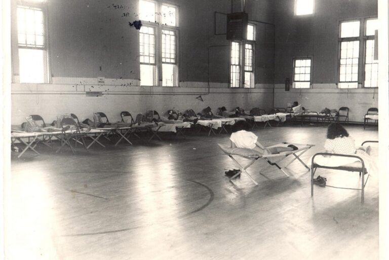 A gymnasium with rows of cots and chairs lined up along the walls, a few people resting on them.