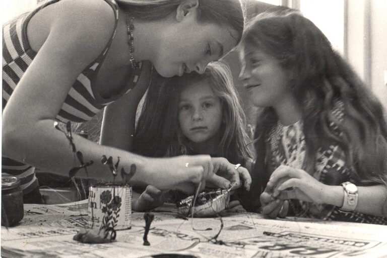 Three girls at a table doing crafts together; one girl focuses on the project, while the others watch and smile.