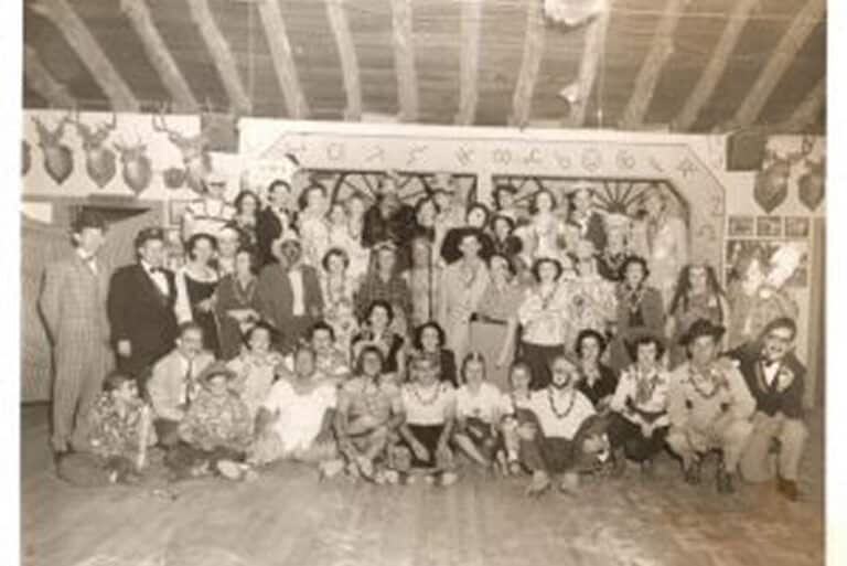 A large group of people in vintage clothing pose together indoors, with animal heads mounted on the walls behind them.
