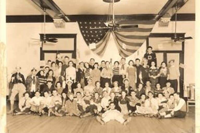 Vintage group photo of people posing in a large room with a draped American flag in the background.