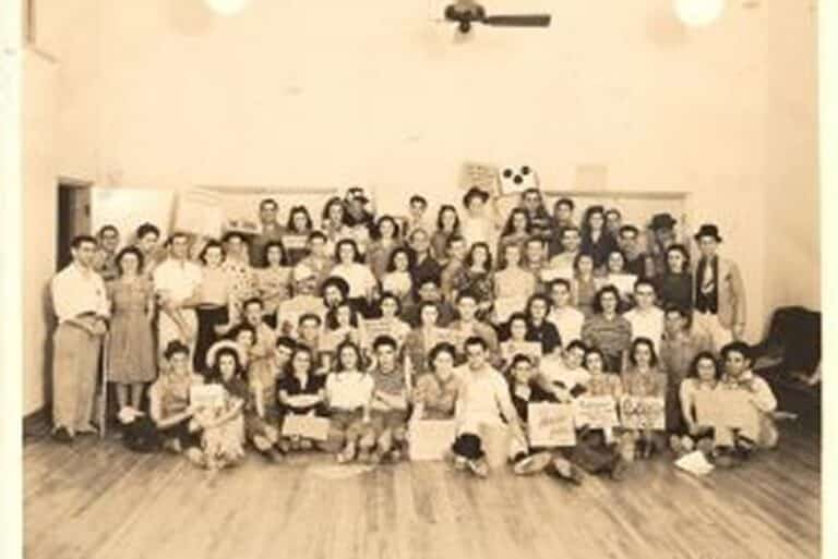 A large group of people posing indoors on a wooden floor, many holding signs, with a panda drawing in the background.