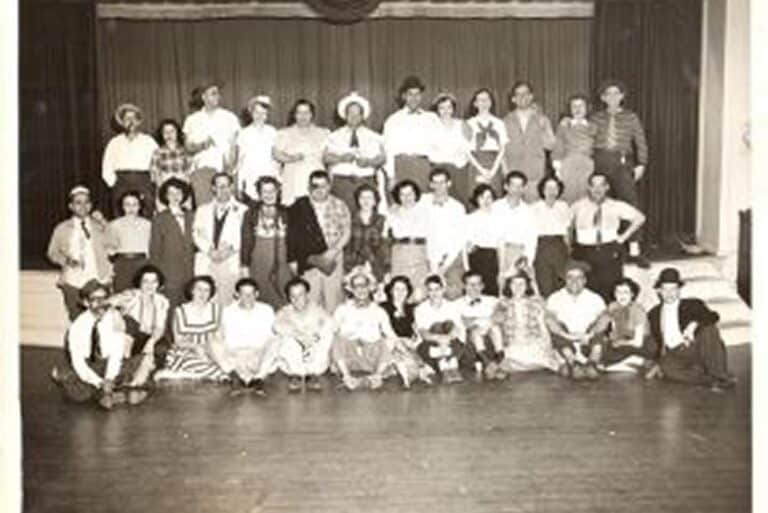 A large group of people in vintage clothing pose together on a stage with curtains in the background.