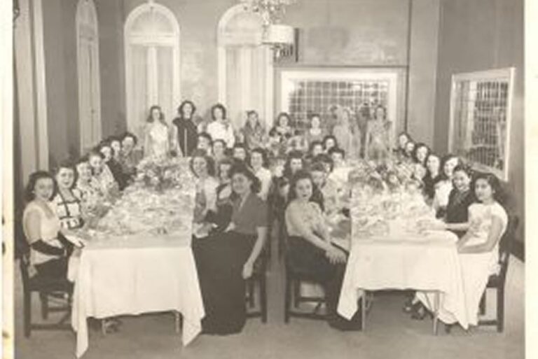 A group of women sit around two long dining tables in a formal room, posing for a black-and-white photograph.