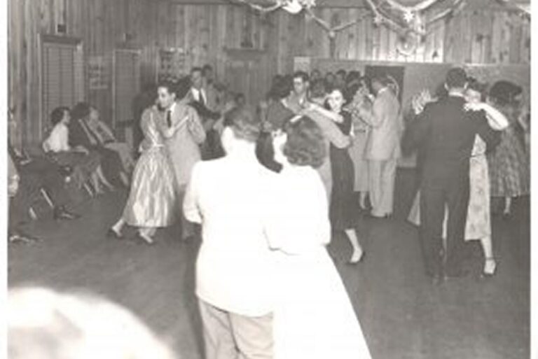 People in formal attire dance in pairs in a wood-paneled room, while others watch from chairs along the wall.