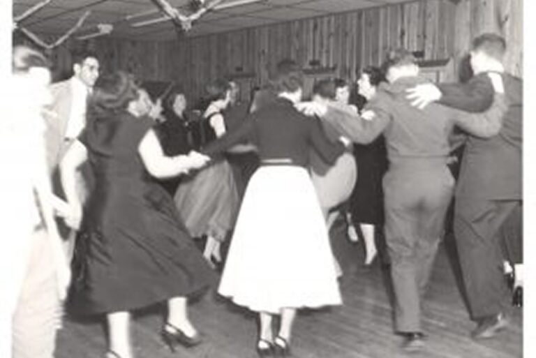 People in vintage clothing dance in a circle in a wood-paneled room, holding hands and smiling.