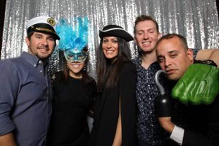 Five people in costumes pose together in front of a silver sequin backdrop, smiling at the camera.