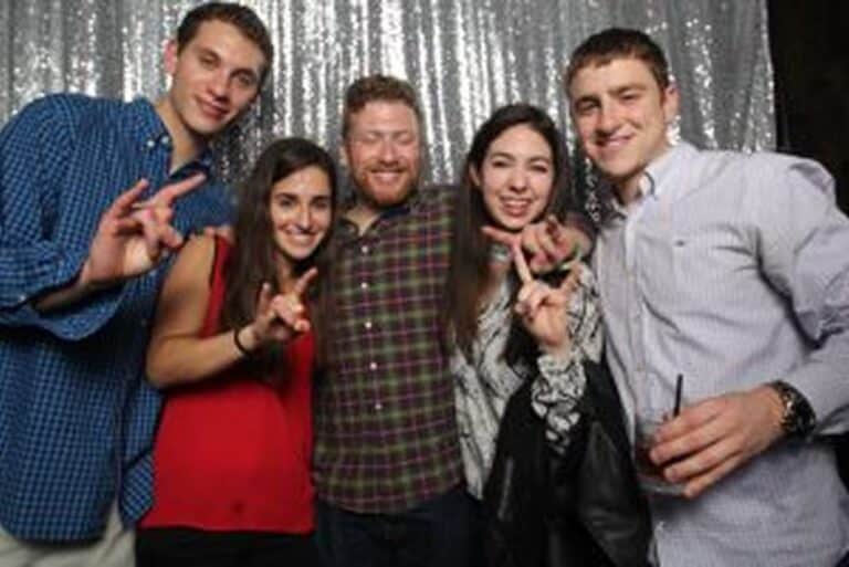 Five people smiling and posing together in front of a silver sequin backdrop at a party.