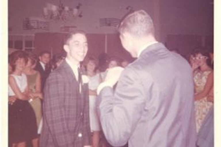 Two men in suits talk at a social gathering, with people standing and chatting in the background.