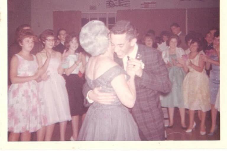 A man and woman dance together at a party while others in formal attire watch and smile in the background.
