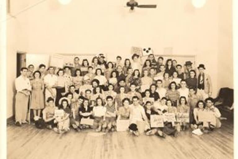 Large group of people pose indoors in vintage clothing, standing and sitting in several rows, smiling at the camera.
