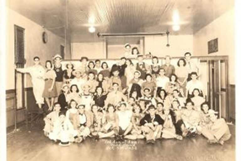 Vintage group photo of people posing in a hall, many wearing costumes and hats, with smiling faces and festive atmosphere.