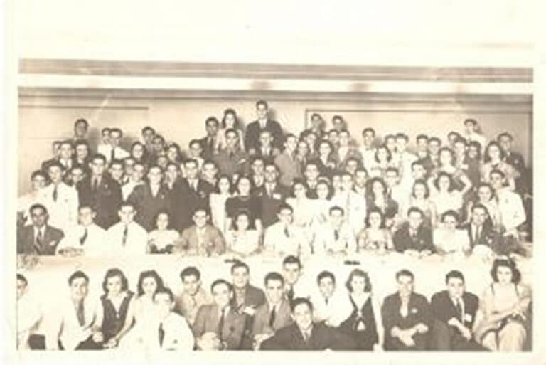 A large group of people in formal attire pose for a vintage black-and-white group photo indoors.