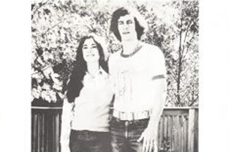 Black and white photo of a woman and man standing outside in front of a wooden fence and trees.