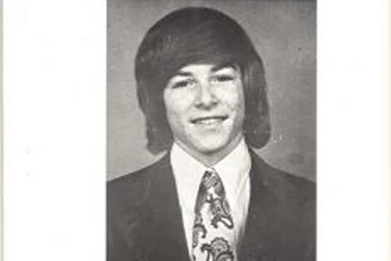 Black-and-white portrait of a young man in a suit and paisley tie with long, side-parted hair, smiling at the camera.